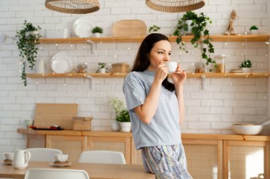 Pijamalı genç bir kadın mutfakta çay içiyor. 