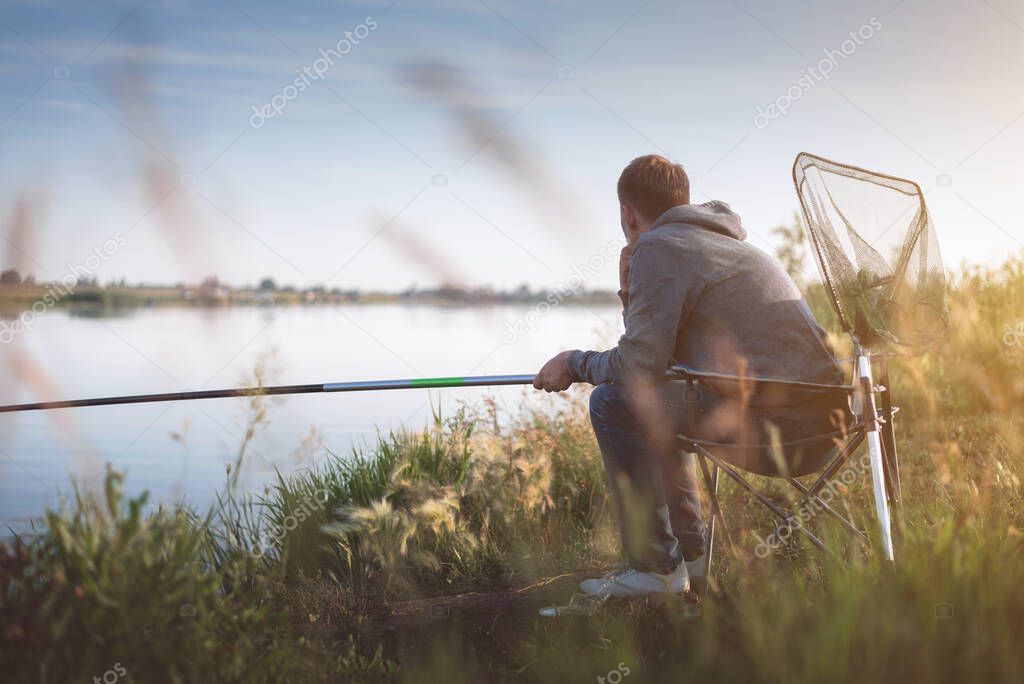 A fisherman with a fishing rod is sitting on chair on the lake branch.