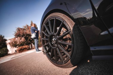Rome, italy 10 september 2025: black sports car wheel and tire on asphalt, low-angle detail shot with person blurred in background under clear blue sky, modern automotive design