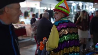 Roma, İtalya, 27 Ekim 2025: Renkli makyajlı ve pullu kostümlü yaşlı bir palyaço kalabalık bir gece panayırında balonları hayvan şekillerine çevirerek insanları eğlendiriyor.