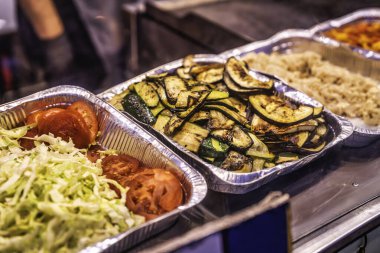 Yemek servisinde alüminyum tepsilerde ızgara kabak, patlıcan dilimleri ve domatesli canlı yeşil salata gibi sağlıklı bir yemeğe hazır çeşitli taze sebzeler yer alıyor.