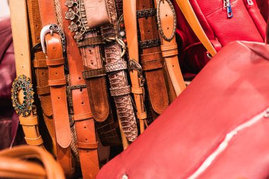 Kırmızı deri çantaların yanında asılı süslü tokaları ve kumaşları olan çeşitli deri kemerler satılık moda aksesuarları sergiliyor.