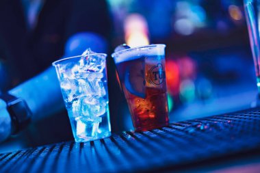 Este, İtalya 28 Ekim 2025: İki buzlu içecek karanlık bir tezgahta bekliyor, parlak mavi kulüp ışıkları ile aydınlatılmış, enerjik bir gece hayatı sahnesi yaratıyor.