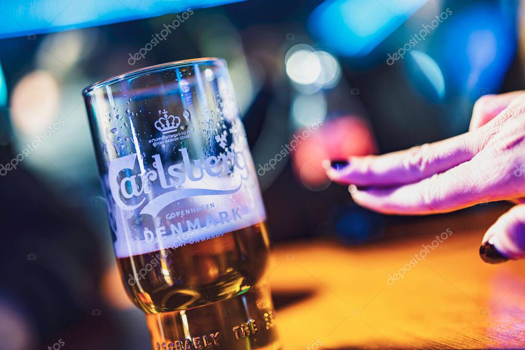 Este, italy 28 october 2025: hand with dark polish touching cold, condensation-covered carlsberg pint on wooden bar counter, evening pub mood and celebration