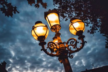 Süslü klasik sokak lambaları karamsar bulutlu bir gökyüzüne karşı duruyor, sıcak bir altın parıltı saçıyor, akşam alacakaranlığı sırasında kentsel çevreye atmosferik aydınlanma sağlıyor.