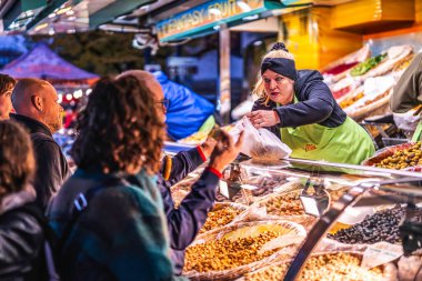 Milan, İtalya, 27 Ekim 2025: satıcı kalabalık bir akşam marketinde zeytin ve sağlıklı atıştırmalıklar sunan bir müşteriye bir paket karışık fındık veriyor.