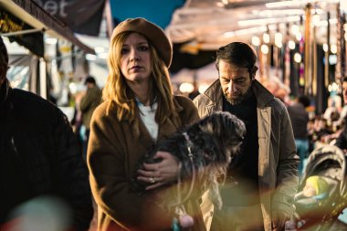 Milan, İtalya 27 Ekim 2025: Köpeğiyle kalabalık bir gece pazarında yürüyen çift, şehir ışıkları ve akşam kalabalığının ortasında sessiz ve dalgın bir şekilde