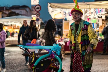 Milan, İtalya, 27 Ekim 2025: Renkli kostümlü ve makyajlı kıdemli palyaço balonu sanatçısı Avrupa 'nın aydınlık bir caddesinde geceleri hayvanlar yapıyor.