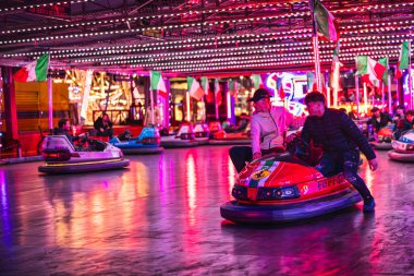 Milan, İtalya 27 Ekim 2025: Neon ışıklarının altında parlayan çarpışan arabaları kullanan iki adam ve eğlenceli bir gece parkında İtalyan bayrakları, birlikte gülüşmeler.