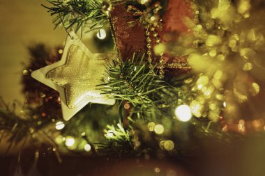 Noel ağacı dalları parlak altın parıltılı bir yıldız süsü ve kırmızı kurdele tutuyor. Bulanık ışıklar ve bokeh efektiyle ılık bir festival atmosferi yaratıyor.