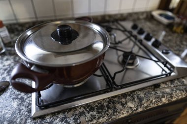 Paslanmaz çelikten kapağı olan kırmızı tencere bir gaz sobasının üzerinde oturuyor. Parlak, modern bir mutfakta yemek hazırlamaya hazır. Ev hayatını ve günlük aktiviteleri simgeliyor.