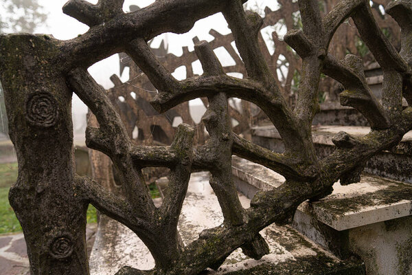Intricate concrete balustrade carved like tree trunks and branches, moss-covered and weathered, forming an ornate, moody staircase handrail in foggy garden ruins