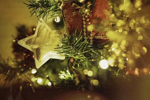 Noel ağacı dalları parlak altın parıltılı bir yıldız süsü ve kırmızı kurdele tutuyor. Bulanık ışıklar ve bokeh efektiyle ılık bir festival atmosferi yaratıyor.