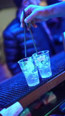 Roma, İtalya, 2 Ocak 2026: Bir barmenin eline yakın plan buz ve tonikli iki plastik bardak eklemek, sonra da onları mavi ışıklı bir gece kulübü partisinde servis etmek