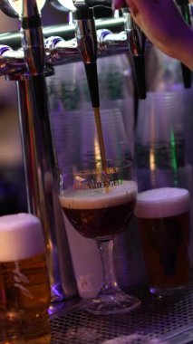 Roma, İtalya, 2 Ocak 2026: Krom musluktan markalı cam kadehe siyah bira döken bir barmene yakın çekim. Bu içecek mükemmel köpüklü bir kafa yaratır.