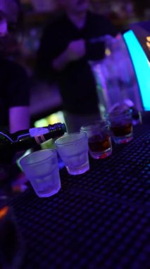 Roma, İtalya 2 Ocak 2026: barmenin eli barın tezgahına bir sıra bardağa kahverengi likör döküyor. Mor neon ışıklar canlı bir parti ortamı yaratıyor.