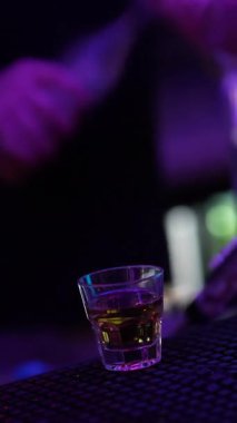 Profesyonel bir barmen, muhtemelen viski ya da burbon gibi alkollü bir içeceği canlı mor ve mavi neon ışıklarla dolu bir bardağa dolduruyor ve dinamik bir gece hayatı atmosferi yaratıyor.