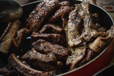 Domuz kaburgası ve yağlı göbek kırmızı bir tavada cızırdıyor, altın sarısı kenarları, otlar ve baharatlar eti kaplıyor, mutfaktaki lezzetli ev yapımı ızgaranın yakın çekimi.