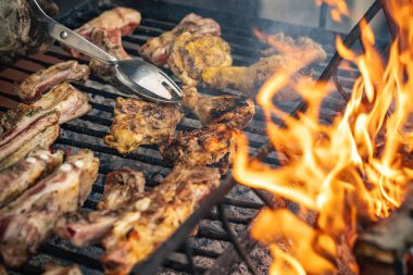 Izgara et ve tavuk sıcak kömür ızgarasında hazırlanıyor, alevler ve maşa kullanan bir el, açık hava yemeklerini ve yaz toplantılarını temsil eden