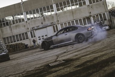 Imola, İtalya 01 Ocak 2026: Gri modifiye edilmiş BMW arabası sürükleniyor, izleyiciler tarafından izlenen endüstriyel binaların yakınındaki bir açık asfalt yüzeyde sigara içiyor.