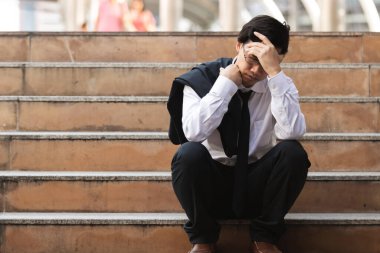 Depresyondaki Asyalı takım elbiseli, elleri başının üstünde merdivenlerde oturan genç bir iş adamı. İşsizlik ve işten çıkarma kavramı.