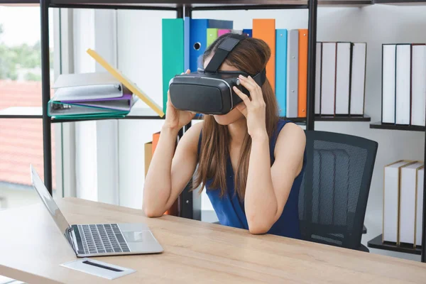 Young Asian woman wearing virtual reality (VR) goggles in workspace. Future technology concept.