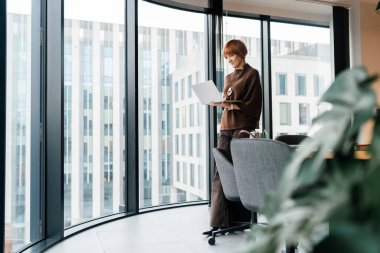 Ofis penceresinin yanında dizüstü bilgisayarla çalışan 50 'li yaşlardaki bağımsız kadın danışmanın tam boy görüşü. Odaklanma, güven, dijital iş akışı, modern ofis, şehir mimarisi, kopyalama alanı