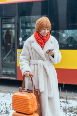 Karlı Avrupa şehrinde bavullu kızıl saçlı kadının akıllı telefon kullanması. Tatil planlama, şehir seyahati, tek başına seyahat, yurt dışına kış gezisi sırasında telefonları kontrol eden kadın gezgin..
