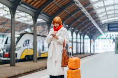 Demiryolu platformunda beyaz önlüklü, bavullu orta yaşlı bir kadın, akıllı telefon programını kontrol ediyor, mesaj veya gecikme bildirimi, dijital bağımlılık, planlama, kontrol, kentsel rutin zaman yönetimi