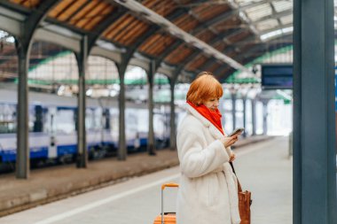 Yetişkin bir kadın, tren istasyonunda bavuluyla beklerken akıllı telefon kullanıyor. Mobil iletişim, kontrol güncellemeleri, toplu taşıma, günlük seyahat yönetimi kavramı.