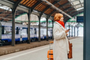 Trenin yanındaki tren istasyonunda bavuluyla bekleyen olgun bir kadın. Kentsel ulaşım, taşınma, toplu taşıma, kış yolculuğu, hareketlilik, günlük hareket, kalkış, yolculuk kavramı.