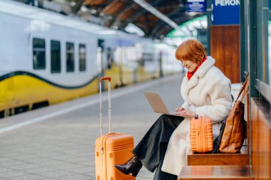 Beyaz saçlı kadın tren istasyonunda tren beklerken dizüstü bilgisayarda çalışıyor. Serbest yaşam tarzı, uzaktan çalışma, hareket kabiliyeti, seyahat, kış yolculuğu boyunca modern çalışma alışkanlığı.