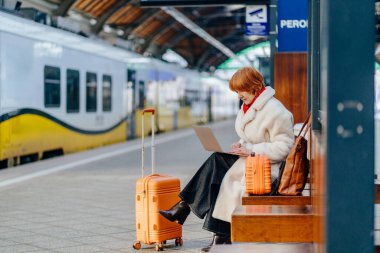 Yetişkin beyaz kadın, tren istasyonunda elinde bavul ve dizüstü bilgisayarla oturuyor. Uzaktan çalışma, esnek program, dijital göçebe zihniyeti, toplu taşıma, kış yolculuğu, iş geçişi.