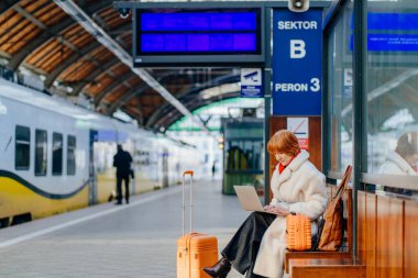 Kendine güvenen orta yaşlı kızıl saçlı kadın tren istasyonunda dizüstü bilgisayar kullanıyor. Kış kenti taşımacılığında ulaşım ve sorumluluk konusunda tecrübeli..