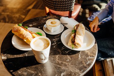 Kahvaltı sırasında iki kahve fincanı ve taze sandviçli mermer kafe masası. Şehir kahve kültürü, günlük yemek, güneş ışığı, modern kafe iç mekan, günlük yaşam tarzı yemek sahnesi.