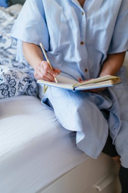 Dikey yakın çekim, tanınmayan orta yaşlı beyaz kadın kalem tutuyor, açık mavi defterle yatakta yazıyor, keten gömlekli elbise, rahat yatak odası tekstili, kişisel planlama, farkındalık pratiği, sessiz bir an..