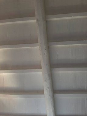 Detail of a ceiling with wooden planks and exposed beams, all painted white, creating a clean and bright atmosphere. 