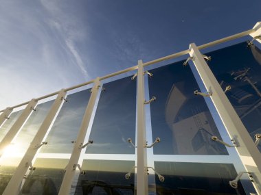 Bir yolcu gemisinin güvertesindeki modern cam bariyerlere bakış açısı, okyanusun üzerindeki batan güneşi yansıtıyor.. 