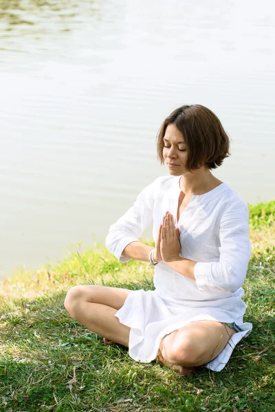 Mulher meditando em pose fácil na grama na margem do rio. O rio está no fundo. — Fotografia de Stock