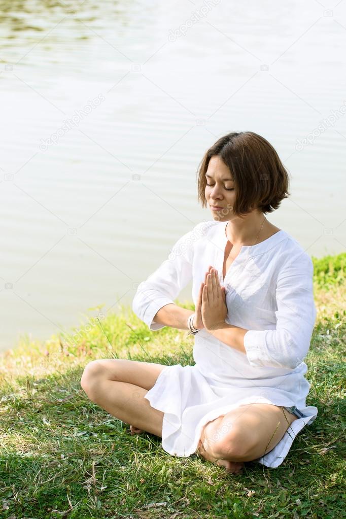 Woman meditating in easy pose on the grass at the river-bank. The river is on the background