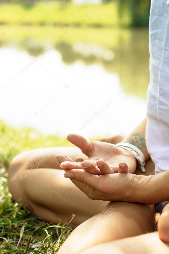 Crossed arms are put on the legs of the girl sitting in meditation pose at the river