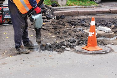 Yansıtıcı kıyafetler giymiş bir yol işçisi eski asfaltı yoldan çıkarmak için elektrikli matkap kullanıyor ve benzin jeneratörü kullanıyor..