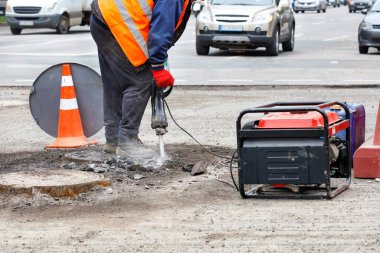 Yansıtıcı giysiler giymiş bir yol işçisi, kanalizasyonun yakınındaki bir yolu elektrikli matkapla tamir ediyor, arka plan olarak bulanık arabaları geçiyor, boşluğu kopyalıyor..