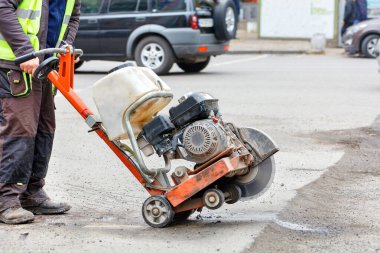 Yansıtıcı giysili bir yol işçisi, yoldaki eski kötü asfaltı kesmek için çalışan elmas testereli bir benzin kesicisini hareket ettirir. Boşluğu kopyala.