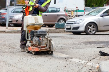 Yansıtıcı giysili bir yol işçisi yol kenarındaki eski asfaltı mazgallı bir caddenin arka planına benzinle kesiyor. Boşluğu kopyala.