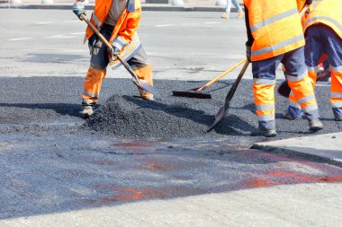 A working group of road workers in orange overalls renews a section of the road with fresh hot asphalt and levels it with shovels and metal slats. clipart