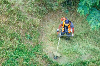 A utility worker in reflective clothing mows tall green grass with a petrol trimmer in circular motions. Copy space, top view. clipart