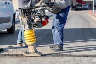 Yol işçileri, parlak güneşli bir günde, seçici bir odak noktasında yolun küçük bir bölümünü asfaltlamak için bir petrol titreşimcisi kullanırlar..