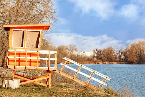 A lonely wooden lifeguard house on the river bank is illuminated by the rays of the bright sun against the background of a blue sky and urban development on the horizon.