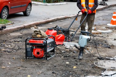 Yansıtıcı turuncu yelek giyen bir yol işçisi, kırmızı konilerle çevrili bir yolda sıkıştırıcı bir plaka, elektrikli matkap ve bir benzin jeneratörü kullanarak kanalizasyon onarıp monte eder. Boşluğu kopyala.